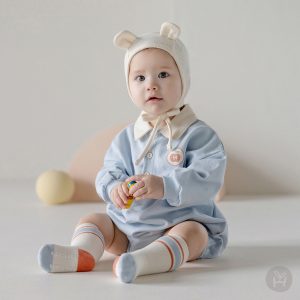 A baby in a light blue Gid coverall with a white and beige hat and matching socks, looking comfortable and stylish