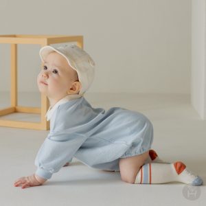 A baby in a light blue Gid coverall with a white and beige hat and matching socks, looking comfortable and stylish